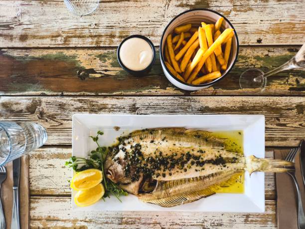 Horizontal high angle photo of a fried whole Sole fish with lemon wedges, caper butter sauce and pea shoot garnish served on a white rectangular ceramic plate with a bowl of fried potato chips and a bowl of garlic mayonnaise on the side, on a repurposed rustic wooden plank table in a fine dining seafood restaurant. Byron Bay, north coast NSW.