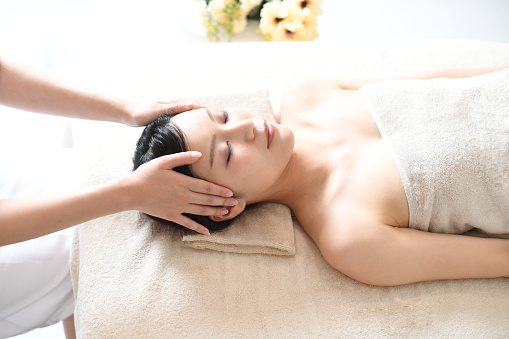 An esthetician working on a young Asian woman lying on her back on a bed receiving a head massage at a spa