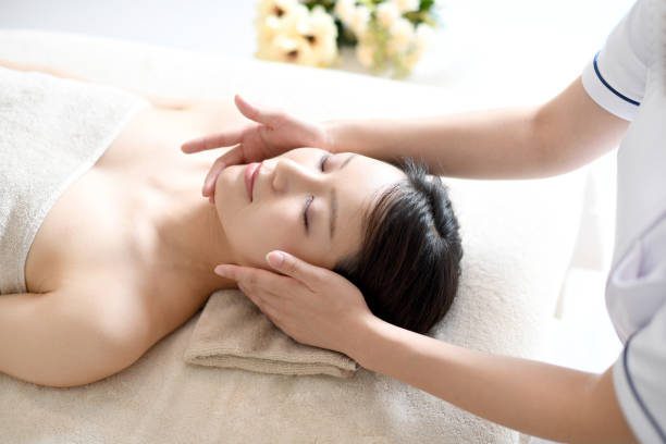 una esteticista que atiende a una joven asiática acostada boca arriba en una cama recibiendo un masaje facial en un spa - tratamiento-de-spa fotografías e imágenes de stock