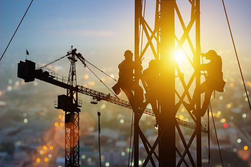 Silhouette of Engineer and worker working crane on steel structures industrial project at building site background, construction site at sunset in evening. Infrastructure, High-rise building Industry