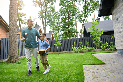 Man and a boy are walking along the green grass near the house, the boy is wearing a baseball glove