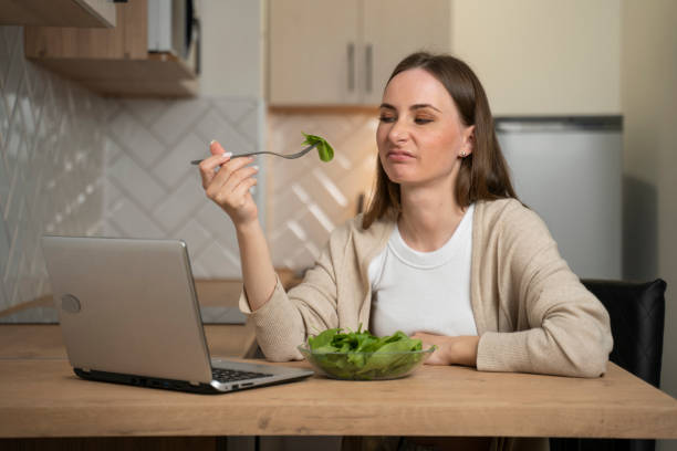 une jeune femme réfléchie mange une salade et manque d’appétit alors qu’elle est assise dans la cuisine et utilise un ordinateur portable. problèmes digestifs, ainsi que des aliments avariés et insipides - overweight women salad frustration photos et images de collection