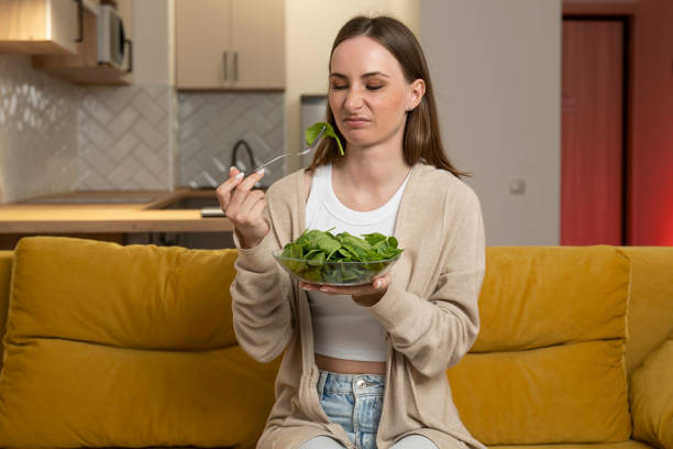 une jeune femme réfléchie mange une salade et manque d’appétit alors qu’elle est assise sur le canapé. problèmes digestifs, ainsi que des aliments avariés et insipides - overweight women salad frustration photos et images de collection
