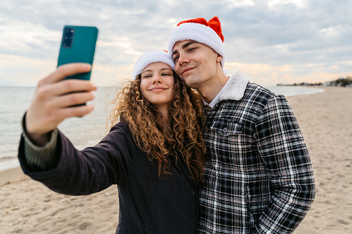 Young couple taking selfies on the beach, wearing Santa hats and celebrating Christmas together in Greece.