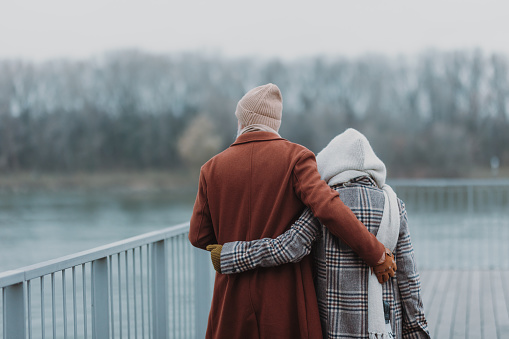 Rear view of elegant senior couple walking near a river, during cold winter day.