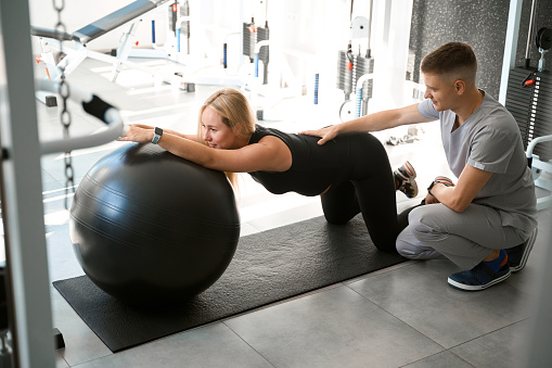 Expectant mother performs relaxing exercises for her back with a fitball, she is helped by a specialist