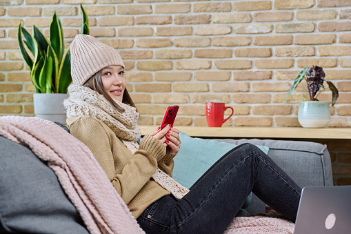 Cold season autumn winter, young woman in cozy woolen sweater hat knitted scarf sitting at home on sofa, in living room, using smartphone. Winter cold holidays, relax, comfort, coziness, lifestyle