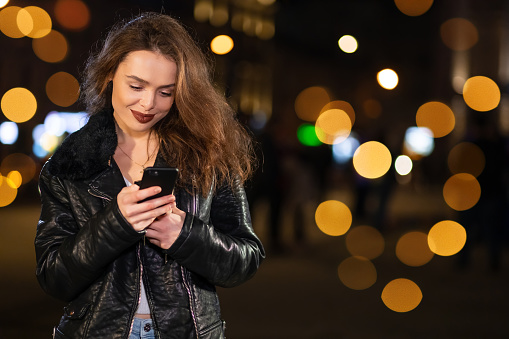 Cheerful brunette woman in stylish outfit enjoying application on smartphone for making online playlist while strolling at evening downtown.
