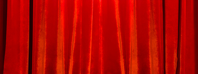 Closed red theatre curtains. Light from a spotlight falls on curved, bright, shiny, textile curtain backdrop. Image for presentation with copy space.