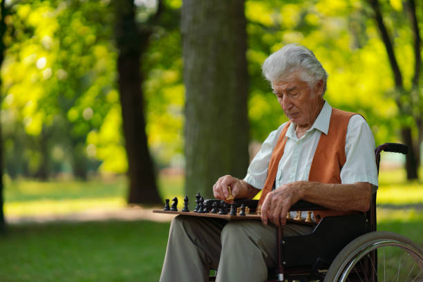 벤치에 체스를 두고 휠체어를 탄 노인이 도시 공원에 혼자 앉아 외로움을 느낀다. - arthritis senior adult rheumatoid arthritis sadness 뉴스 사진 이미지