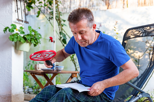 Man Outdoors Having Summer Afternoon Break Reading Book, Enjoy Coffee and Try to Catch Mosquito with Electric Flyswatter