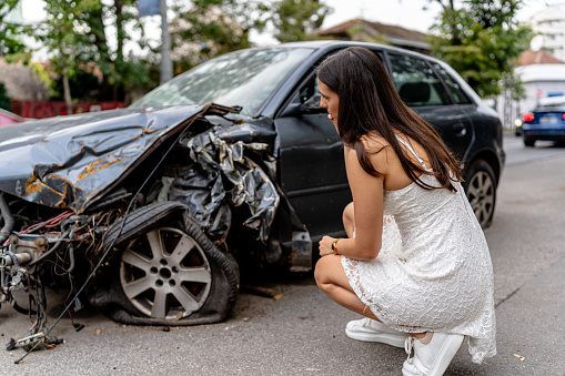 Post-collision, a young woman stands beside her car, attentively examining the extent of the damage to comprehend the aftermath