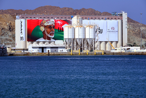 Mutrah, Muscat, Oman: cereals and cement pier at Port Sultan Qaboos - the cement silos are operated by Raysut Cement  - grain silos with large canvas with Sultan Qaboos and the Omani flag. Omran and DAMAC Properties logo on the left side. Al Hajar mountains in the background.
