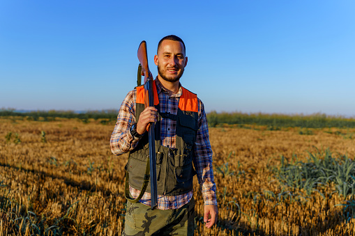 Young Caucasian male hunter cheerfully standing in a field on a sunny day, while holding a rifle in his hands. He is smiling and looking at the camera.