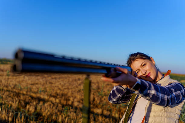Young female hunter standing in a wheat field, while holding a shotgun in her hands. The focus is on her while she is aiming at her prey.
