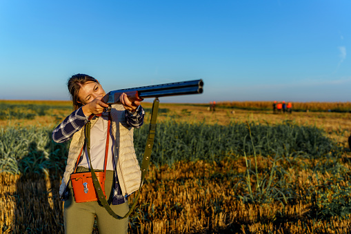 Young Caucasian female hunter holding a shotgun, while standing in a field and pointing at the direction of her prey.