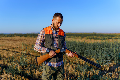 Young male Caucasian hunter checking the bullets in his shotgun, while holding it with both hands. He is standing in a wheat field.
