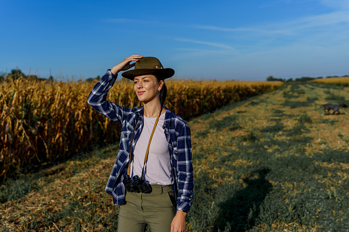 Young Caucasian woman adjusting her hat while standing in nature on a sunny morning. She has binoculars around her neck.