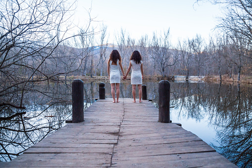 Back view of twin sisters in same white dresses holding hands while standing on wooden pier at calm lake on autumn day