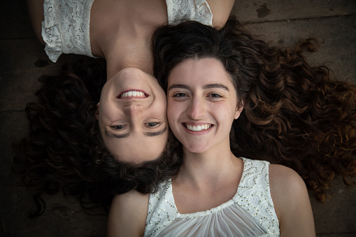From above of young and beautiful twin sisters in white dresses lying opposite on wooden floor smiling at camera
