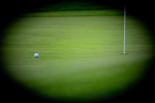 A golf ball comes to rest a few feet from the hole with a flag pole in it. The ball is drawn out by selective focus. The image is vignetted.