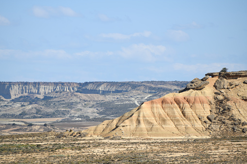 The photo shows a rocky slope with strata showing and signs of erosion. In the distance there are further slopes, hills and erosion. All around are signs of eroded rock. The sky is blue.\n\nThe photo is taken in the Bardenas Reales (sometimes referred as Bárdenas Reales) national park. It is a semi-desert natural region, or badlands, of some 42,000 hectares (420 km2; 104,000 acres) in southeast Navarre, bordering Aragon, Spain. It is located in the middle of the depression of the Ebro valley, at the foot of the mountains of the Yugo and the Zaragoza region of Cinco Villas, and is a UNESCO Biosphere Reserve.  It comprises of the natural Reserve ‘Rincón del Bú’ and the Reserva Natural ‘Caídas de la Negra’ .The photo was taken in May2023