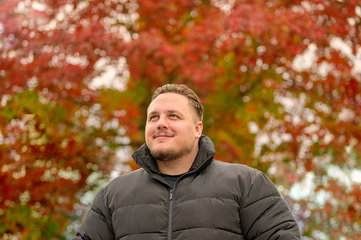 Close up portrait of a young man in a thick winter jacket looking happily to the side, trees in autumnal colors in the background