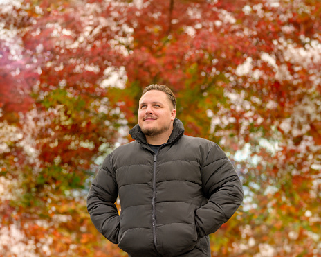 Upper body portrait of a young man in a thick winter jacket looking happily to the side, trees in autumnal colors in the background