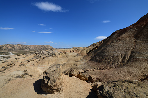 The photo shows a rocky slope with signs of erosion. In the distance there are further rock formations, and mesa. All around are signs of eroded rock. The sky is blue.\n\nThe photo is taken in the Bardenas Reales (sometimes referred as Bárdenas Reales) national park. It is a semi-desert natural region, or badlands, of some 42,000 hectares (420 km2; 104,000 acres) in southeast Navarre, bordering Aragon, Spain. It is located in the middle of the depression of the Ebro valley, at the foot of the mountains of the Yugo and the Zaragoza region of Cinco Villas, and is a UNESCO Biosphere Reserve.  It comprises of the natural Reserve ‘Rincón del Bú’ and the Reserva Natural ‘Caídas de la Negra’ .The photo was taken in May2023