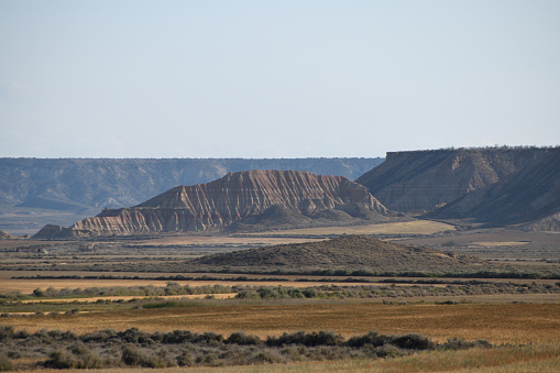 The photo shows the badlands of Bardenas Reales National Park. The erosion is clearly shown. In the distance is a big plateau and in front of it a mesa. The sky is blue.\n\nThe photo is taken in the Bardenas Reales (sometimes referred as Bárdenas Reales) national park. It is a semi-desert natural region, or badlands, of some 42,000 hectares (420 km2; 104,000 acres) in southeast Navarre, bordering Aragon, Spain. It is located in the middle of the depression of the Ebro valley, at the foot of the mountains of the Yugo and the Zaragoza region of Cinco Villas, and is a UNESCO Biosphere Reserve.  It comprises of the natural Reserve ‘Rincón del Bú’ and the Reserva Natural ‘Caídas de la Negra’ .The photo was taken in May2023