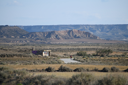 The photo shows the badlands with a plateau in the distance and a blue sky. There is a derelict building in the middle left of the photo and a distant road with cyclist on the left. A mesa in the distance shows sign of rutts from erosion.

The photo is taken in the Bardenas Reales (sometimes referred as Bárdenas Reales) national park. It is a semi-desert natural region, or badlands, of some 42,000 hectares (420 km2; 104,000 acres) in southeast Navarre, bordering Aragon, Spain. It is located in the middle of the depression of the Ebro valley, at the foot of the mountains of the Yugo and the Zaragoza region of Cinco Villas, and is a UNESCO Biosphere Reserve.  It comprises of the natural Reserve ‘Rincón del Bú’ and the Reserva Natural ‘Caídas de la Negra’ .The photo was taken in May2023