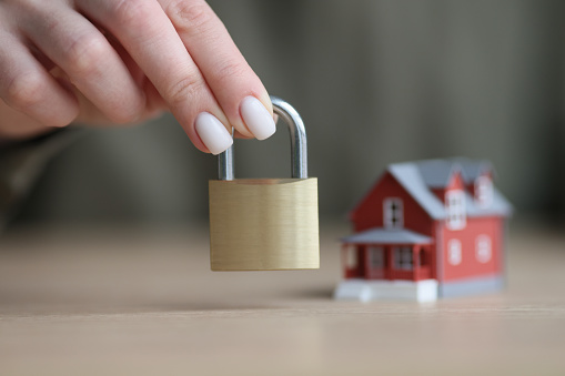 Hand of woman holding iron door lock against blurred small model of cottage house. Two-story building on wooden table. Home security and insurance concept