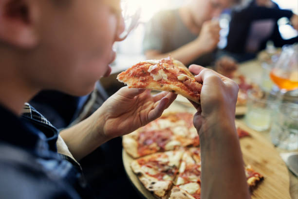 Teenagers have ordered a pizza for lunch and they are enjoying eating pizza.Closeup of the pizza on the wooden pizza plate. Hands reaching for pizza slices.Shot With Canon R5
