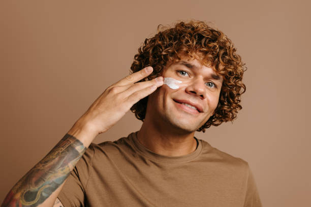 Handsome young man taking care of skin while applying cosmetic cream on face while standing on brown background