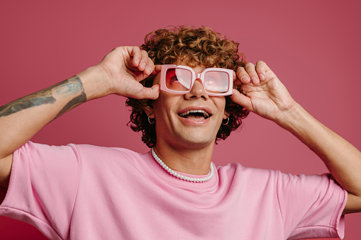 Fashionable young curly hair man adjusting his retro styled eyeglasses and smiling on pink background