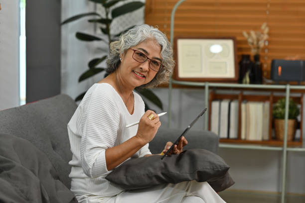 istock Senior Asian woman using a digital tablet while sitting on the sofa, looking at camera 1735416324