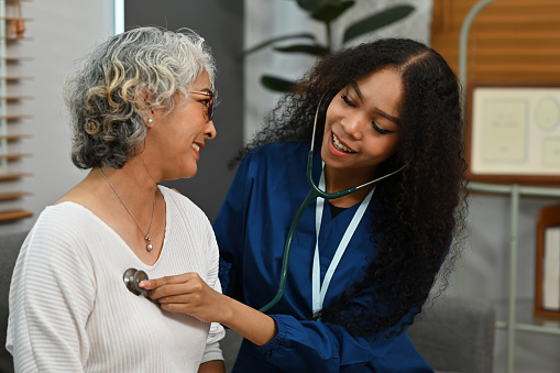 An African-Asian doctor wearing a scrub uniform listening to an older woman lung or heart sounds with a stethoscope during a visit at home, medical checkup and health care concept.
