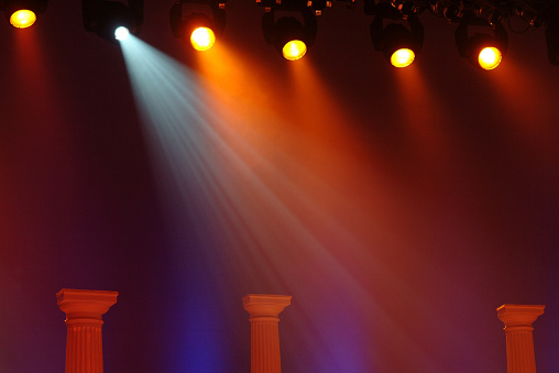 Approximately six blue and yellow stage spot lights shinning from above the stage. The lights are hanging and the frame supporting them is slightly visible. The stage lights are pointed downward toward three Greek columns. There is fog in the air creating richer tones from the lights. 
