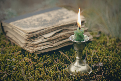 Old battered magic book and a burning candle on the green moss in the forest, close up, selective focus. Vintage, fairy tale, novel cover, witchcraft, mystery, victorian Gothic concept