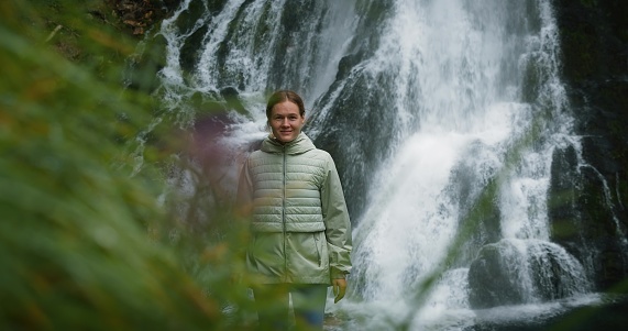Woman traveler enjoying waterfall in the highlands of Austria. Traveling in the mountains, adventure and trip concept. Girl contemplates the beauty of nature in the forest.