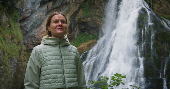 Woman enjoying a mighty waterfall in forest at rainy day. Girl takes a deep breath of fresh air in autumn nature. Gollinger Wasserfall in Austria. Hiker contemplates the beauty of water.