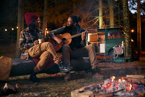 Happy African American man playing acoustic guitar while relaxing with his girlfriend by bonfire at camper trailer park at night.