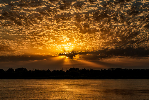 Evening dawn, the clouds are painted in bright rich orange and yellow colors by the sun behind the clouds. Beautiful sun rays of light emanate from behind the clouds. There is a light path on the surface of the water.
