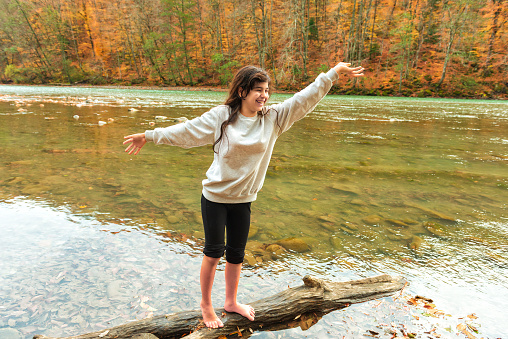 a joyful girl stands on a log lying in the water in the river. Deep autumn, cool weather, bright orange leaves on the trees.