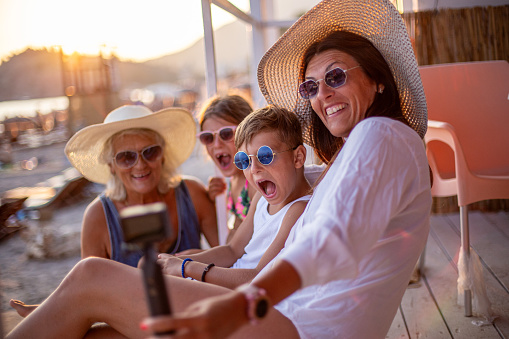 Grandmother, mother and children taking selfie with action camera on beach with hats, sunglasses