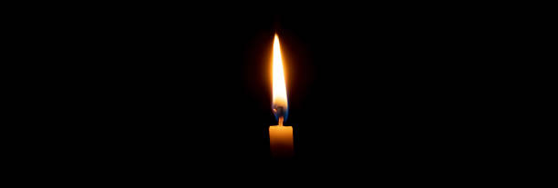 istock Single burning candle flame or light glowing on a small yellow candle on black or dark background on table in church for Christmas, funeral or memorial service with copy space. 1735530047