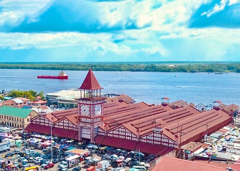 This image shows our main port and market area with a commercial ship commuting the astonishing Demerara River set as the backdrop.
   The Stabroek Market is the largest market in Georgetown, Guyana. 
Located in the center of the capital city, the market is housed in an iron and steel structure with a prominent and Iconic clock tower designed by an American engineer Nathaniel McKay, and constructed by the Edgemoor Iron Company of Delaware, USA over the period 1880-1881.