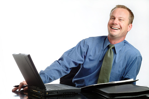 Horizontal photograph of a handsome middle aged caucasian businessman sitting at his desk. He has his laptop and paperwork in front of him. He is looking off camera slightly with a big smile and maybe a little laugh. The image was shot in the studio on a white background. 