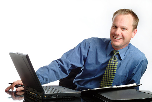 Horizontal photograph of a handsome middle aged caucasian businessman sitting at his desk. He has his laptop and paperwork in front of him. He is making eye contact with the camera and has a smile. The image was shot in the studio on a white background. 
