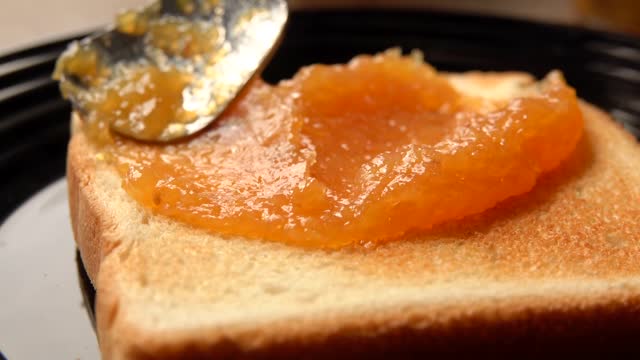 Close-up view of the spoon spreading cooked orange jam on the toasted bread